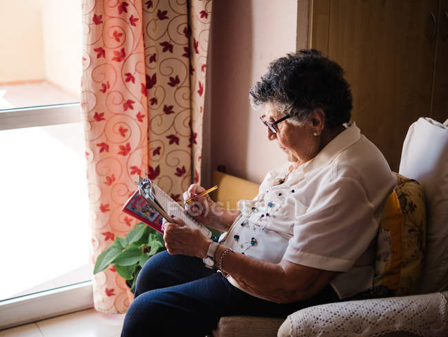Вид сбоку пожилой женщины в блузке и очках, сидящей на кресле у окна и решающей кроссворды дома — стоковое фото