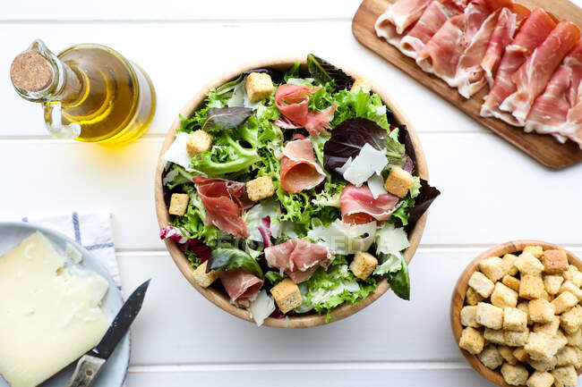 From above bowl of delicious lettuce salad with bacon and croutons placed on white tabletop near bottle of oil and slice of cheese — Stock Photo