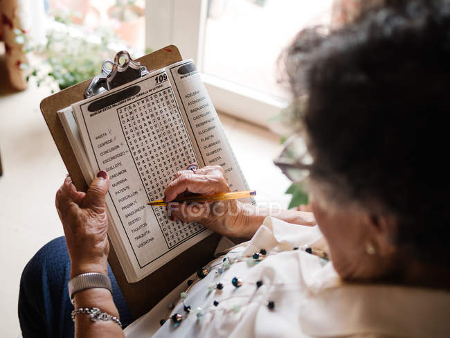 Side view of senior woman in blouse and glasses sitting on armchair near window and solving crosswords at home — Stock Photo