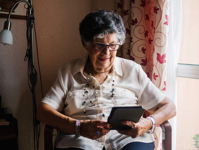 Senior woman in shirt and glasses sitting on armchair and reading e-book in apartment — Stock Photo