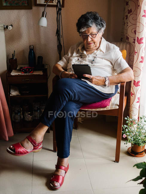 Пожилая женщина в рубашке и очках сидит на кресле и читает электронную книгу в квартире — стоковое фото