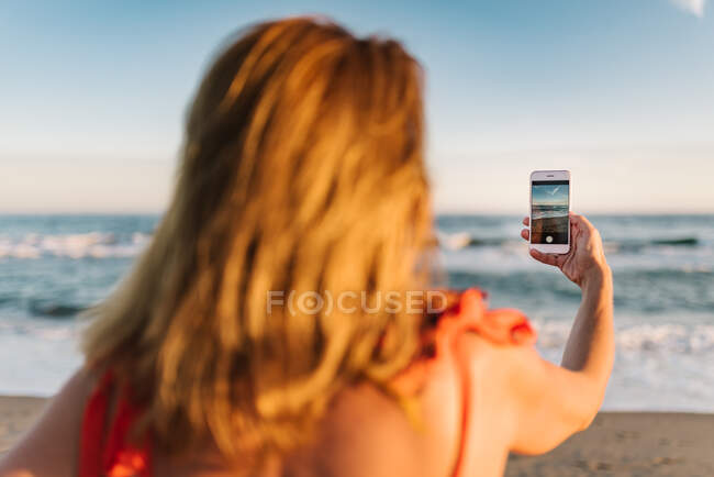 Vista trasera de una mujer irreconocible tomando una foto con teléfono móvil en la playa vacía y arenosa - foto de stock