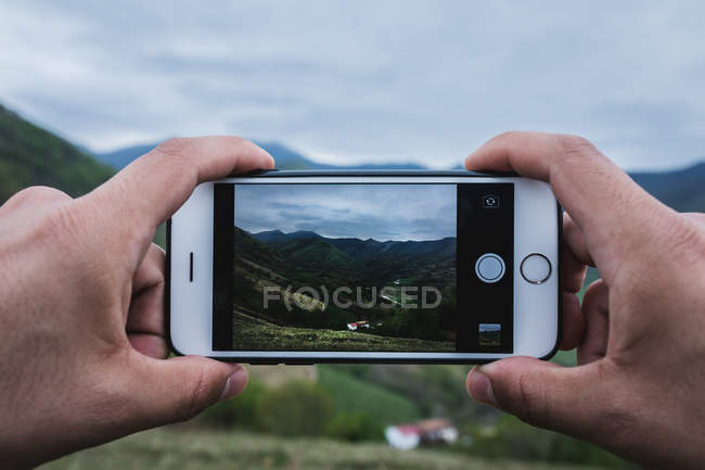 Closeup hands of anonymous traveler using smartphone to take photo of green hilly terrain and village on cloudy day — Stock Photo