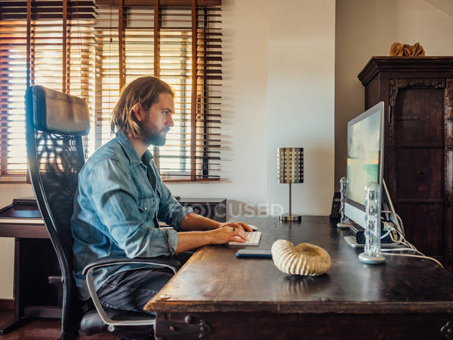 Side view of serious man in denim shirt sitting in comfy armchair while surfing computer at wooden table in home office — Stock Photo