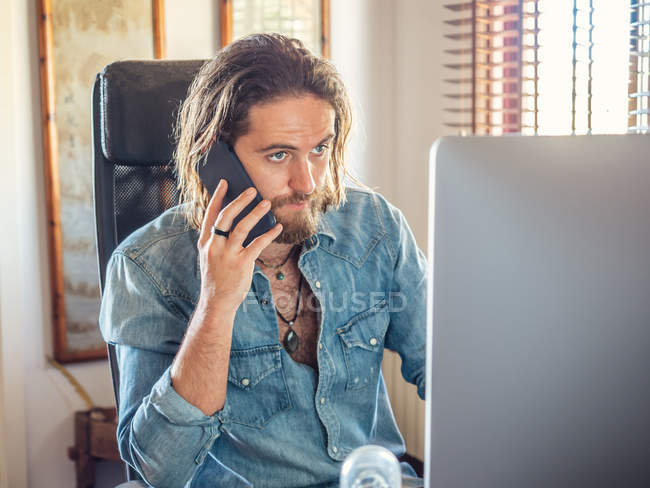 Attentive young man browsing PC and talking on phone in flat — Stock Photo