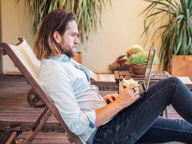 Vista laterale dell'uomo barbuto che mangia mela e utilizza il computer portatile mentre si rilassa sulla sedia a sdraio sul balcone a casa — Foto stock