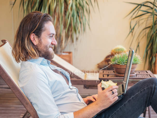 Schöner junger Mann surft Laptop und genießt Apfel auf Lounge in Wohnung — Stockfoto