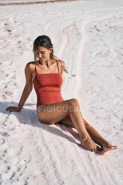 Slender woman in red swimsuit posing on on salty shore of lake looking down in sunshine — Stock Photo