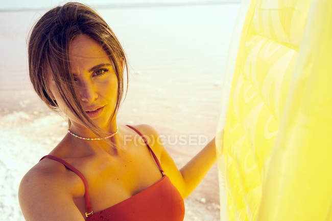 Ніжна жінка в червоному купальнику з яскравим повітряним матрацом, що стоїть на березі моря на сонячному світлі, дивлячись на камеру — стокове фото