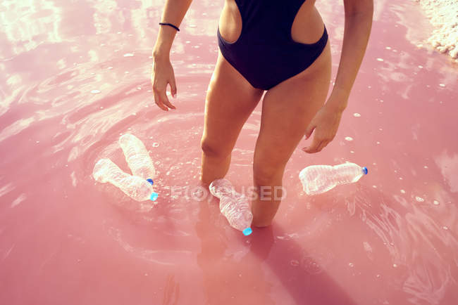 Cropped view of tanned woman in swimwear walking in pink salty water contaminated with plastic bottles at red lagoon in summer — Stock Photo