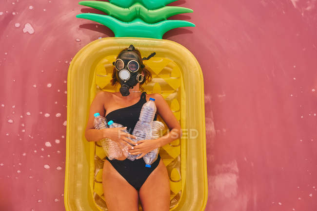 Вид зверху на жінку в респіраторі з пластиковими пляшками, що лежать на ананасовому надувному матраці в червоній воді тропічної лагуни — стокове фото