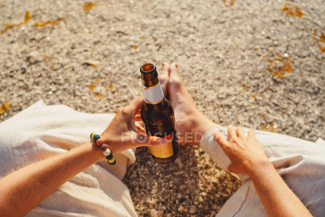 Imagen recortada de la mujer sentada en piedra en la costa y bebiendo cerveza de botella - foto de stock