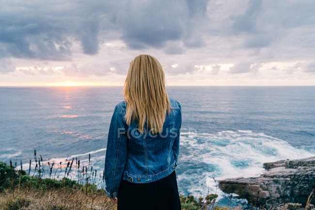 Rückansicht einer blonden Frau, die in Wolken allein am ruhigen Meeresufer chillt und die malerische Landschaft betrachtet — Stockfoto