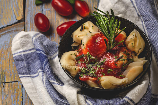 Готовые равиоли с томатным соусом и травами на тарелке рядом с помидорами и ткань на деревянном деревенском столе — стоковое фото