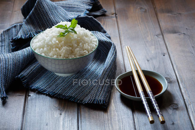 Чаша традиционного японского риса на сером полотенце и палочки для еды на блюдце с соевым соусом на деревянном столе . — стоковое фото