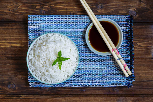 Vue du dessus du bol de riz japonais traditionnel sur serviette bleue et baguettes sur soucoupe avec sauce soja sur table en bois . — Photo de stock