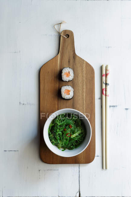 Von oben flaches Sushi mit Algensalat in Schüssel auf Schneidebrett und Essstäbchen auf rustikalem Tisch — Stockfoto