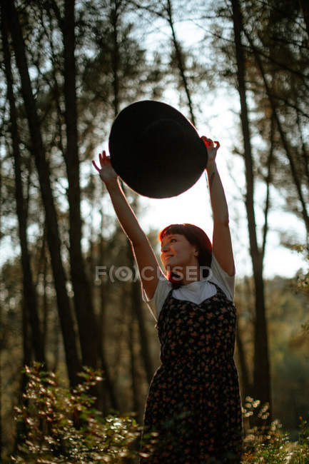 Mujer alegre vestida con ropa retro que sostiene un sombrero caminando sola en el bosque mientras mira hacia otro lado. - foto de stock