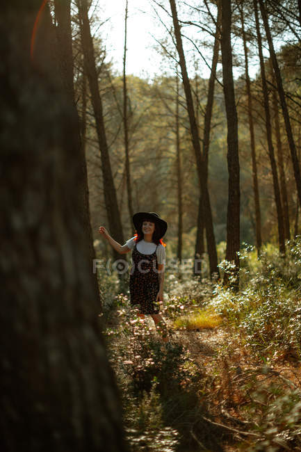 Cheerful woman in hat smiling and walking amidst tall trees on sunny day in green forest — Stock Photo