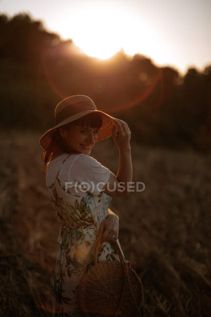 Вид сбоку женщины в ретро-платье и шляпе, идущей по полю к закатному небу, глядя в камеру — стоковое фото