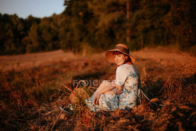 Seitenansicht einer Frau in Retro-Kleid und Hut, die inmitten eines Feldes sitzt und in die Kamera blickt — Stockfoto