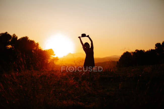 Silhouette of woman raising hands with hat and dancing against bright sunset sky in field — Stock Photo