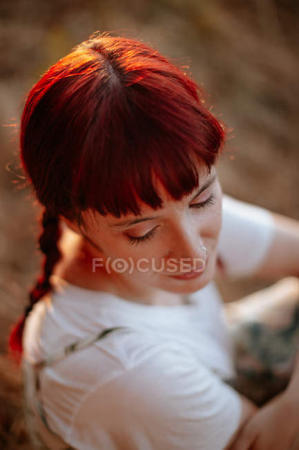 Von oben attraktive Frau mit roten geflochtenen Haaren, die die Augen schließt, während sie abends auf verschwommenem Grund auf dem Feld sitzt — Stockfoto