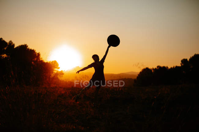 Silhouette of woman raising hands with hat and dancing against bright sunset sky in field. - foto de stock