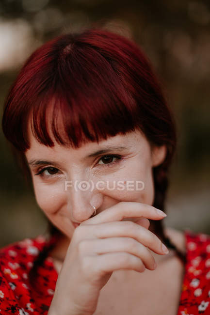 Cheerful woman with red hair giggling and looking at camera while spending time on blurred background of countryside — Stock Photo