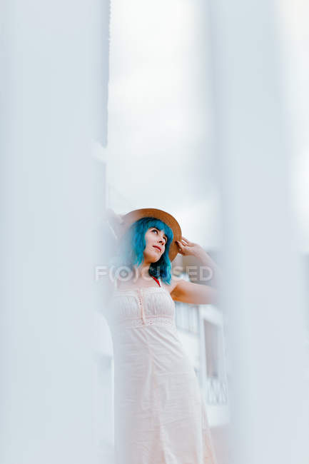 Расслабленная женщина с голубыми волосами в шляпе и сарафане прогуливается по городской улице в летний день — стоковое фото
