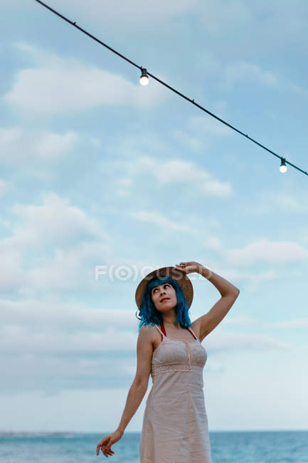 Inspired woman with blue hair in hat and sundress strolling along concrete wharf beside colorful sea — Stock Photo