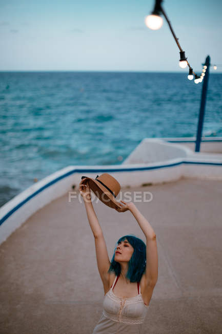 Вдохновлённая женщина с голубыми волосами в шляпе и сарафане прогуливается вдоль бетонной пристани у красочного моря — стоковое фото
