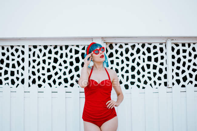 Fashionable woman in bright sunglasses with blue hairstyle in red swimsuit standing with hand aside near design wall — Stock Photo