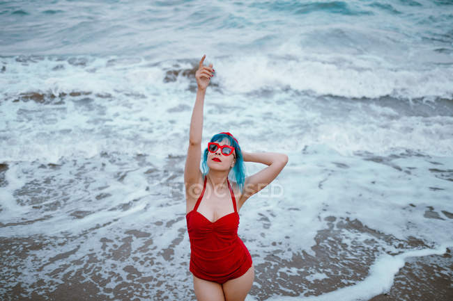 Femme à la mode avec les cheveux bleus en maillot de bain rouge profitant de l'eau restant dans les vagues mousseuses avec les mains levées — Photo de stock