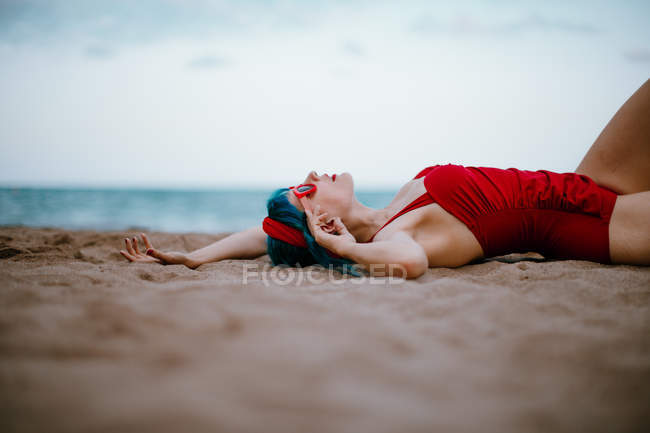 Модная женщина с голубыми волосами в красном ярком купальнике, наслаждающаяся лежащим на песчаном пляже с протянутыми руками — стоковое фото
