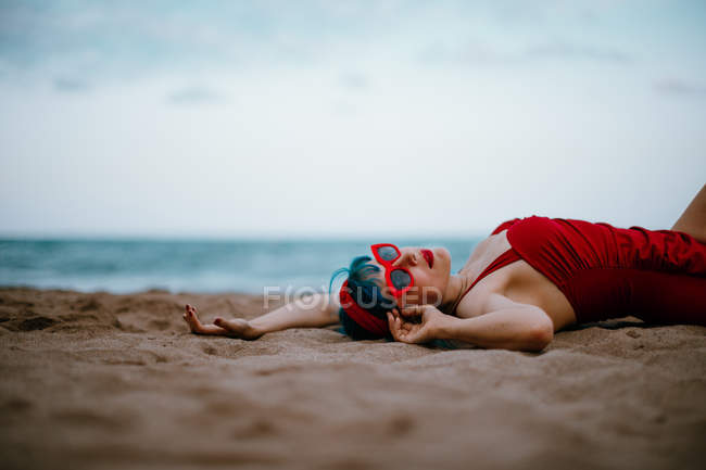 Modische Frau mit blauen Haaren im roten hellen Badeanzug genießt es, mit gestreckten Händen am Sandstrand zu liegen — Stockfoto