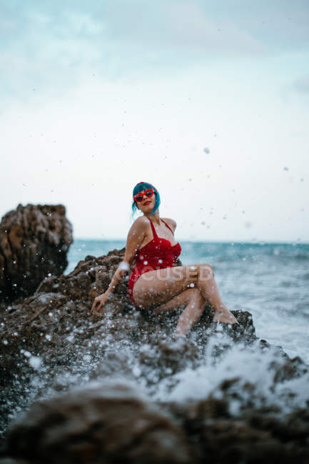 Donna alla moda con capelli blu in costume da bagno rosso brillante che riposa comodamente seduto su pietra rocciosa scura in acqua di mare schiumosa — Foto stock