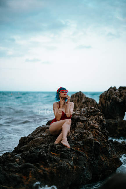 Модная женщина с голубыми волосами в красном ярком купальнике, отдыхающая комфортно, сидя на темном каменистом камне в пенной морской воде — стоковое фото
