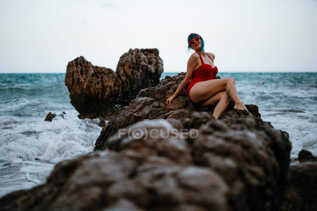 Mujer de moda con pelos azules en traje de baño rojo brillante que descansa cómodamente sentado en piedra rocosa oscura en agua de mar espumosa - foto de stock