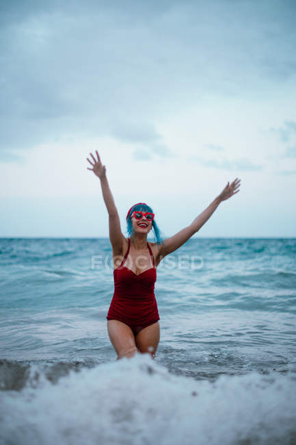 Модна жінка з блакитним волоссям у червоному купальнику насолоджується водою, що перебуває у пінистих хвилях з піднятими руками вгору — стокове фото