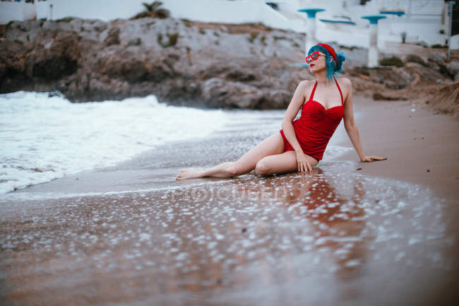 D'en bas femme à la mode avec les cheveux bleus en maillot de bain lumineux rouge profitant assis sur la plage de sable — Photo de stock