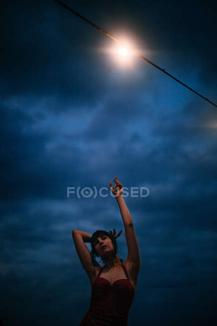 Спокійна жінка в червоному вінтажному купальнику рухається з рукою вгору під темним хмарним небом і яскравим прожектором — стокове фото