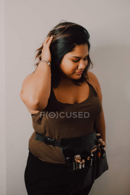 Jovem artista de maquiagem feminina com pincéis avental saco de pé no fundo cinza e ajustar o cabelo — Fotografia de Stock