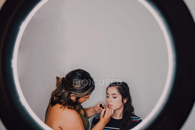 Giovane professionista trucco artista mettendo rossetto sulle labbra dei giovani clienti in studio attraverso un anello di luce — Foto stock