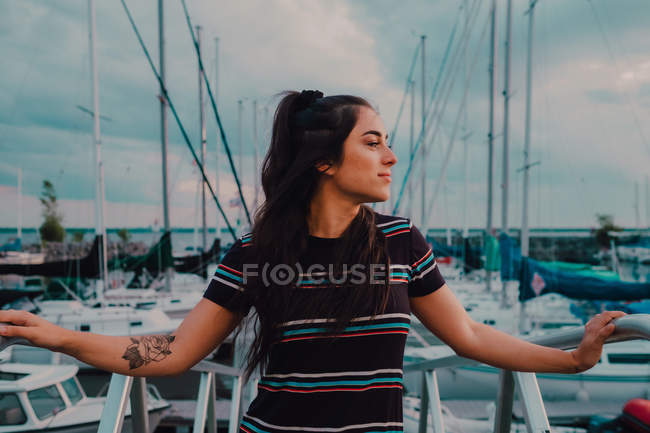 Счастливая молодая татуированная женщина в платье стоит на пристани, заполненной яхтами и лодками — стоковое фото