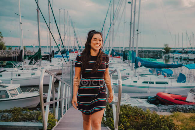 Felice giovane tatuato donna in abito camminando lungo molo pieno di yacht e barche — Foto stock