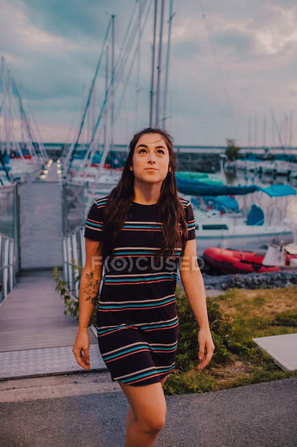 Happy young tattooed woman in dress walking along wharf filled with yachts and boats — Stock Photo