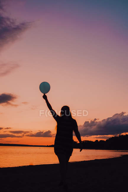 Вид сзади анонимной женщины с воздушным шаром и идущей вдоль берега во время красивого заката — стоковое фото