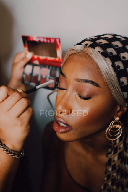 Atractiva mujer negra adulta que aplica sombra ocular del artista profesional de maquillaje en estudio. - foto de stock