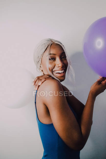 Mulher adulta preta alegre com balões coloridos olhando para a câmera enquanto está em pé no fundo branco — Fotografia de Stock